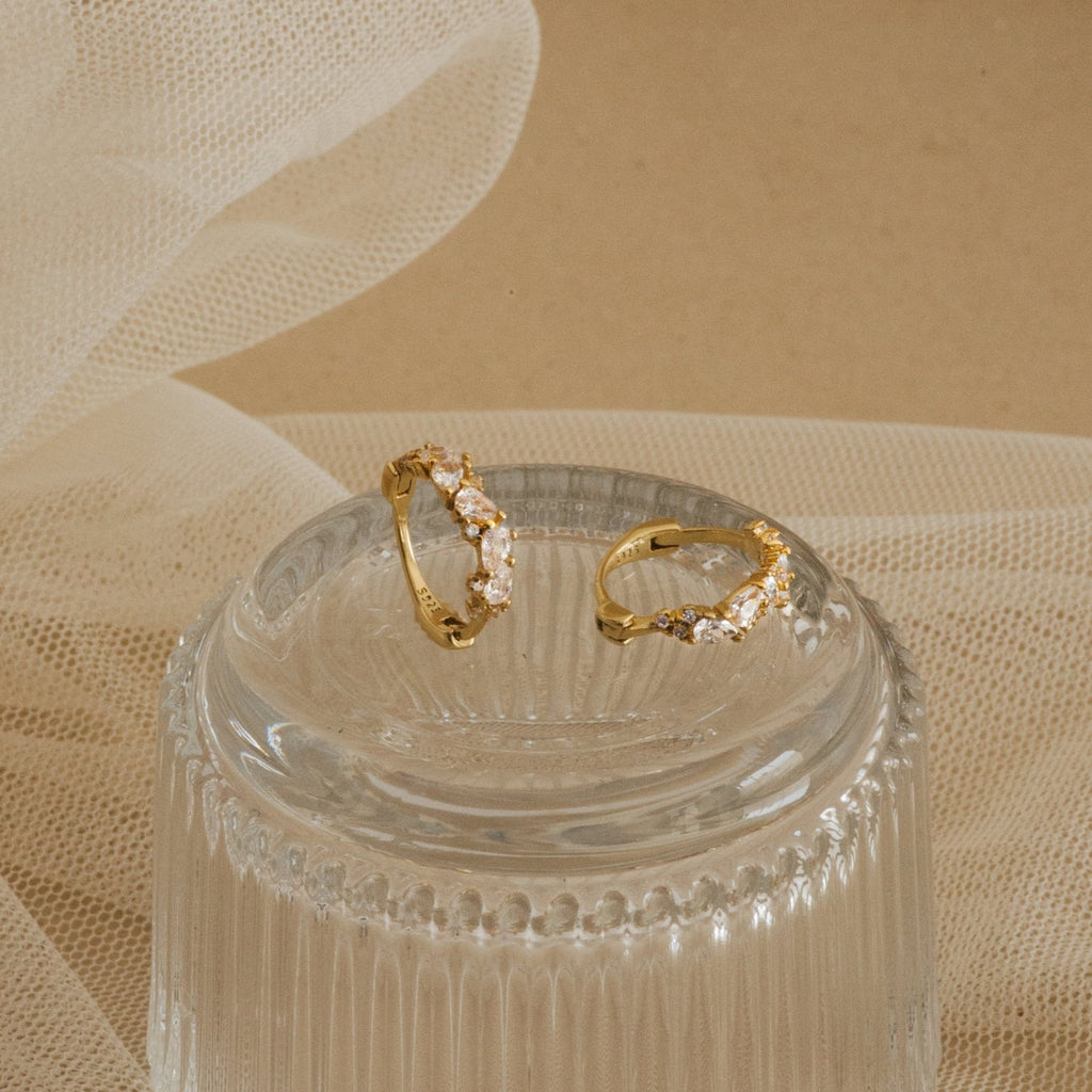 A pair of Sprinkle Diamond Huggies resting on a ribbed glass dish. These small gold hoop earrings are adorned with a scattered arrangement of variously shaped diamond CZ stones, including marquise, round, and oval cuts.