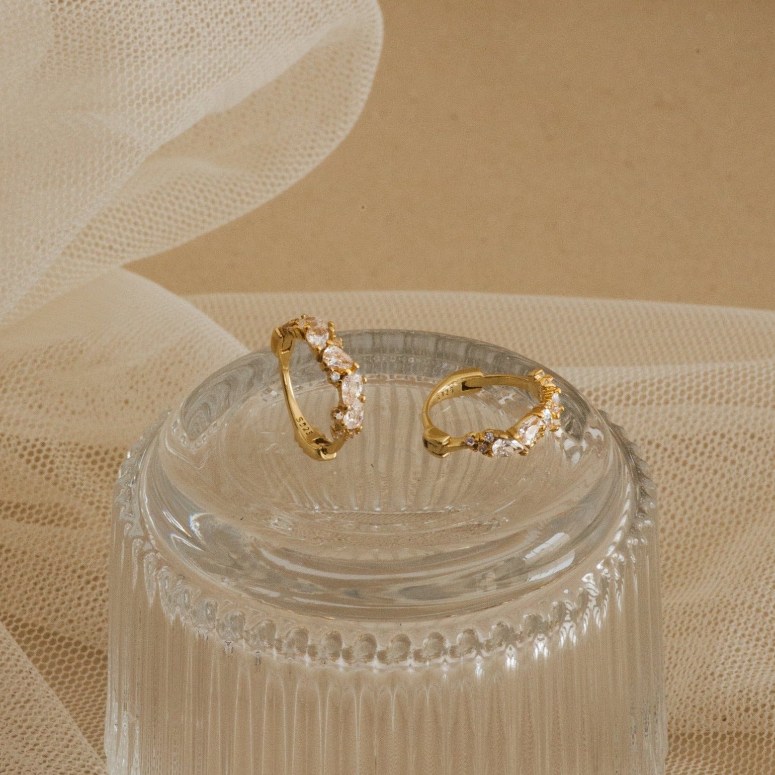 A pair of Sprinkle Diamond Huggies resting on a ribbed glass dish. These small gold hoop earrings are adorned with a scattered arrangement of variously shaped diamond CZ stones, including marquise, round, and oval cuts.
