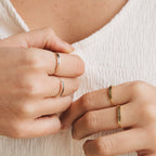 Close-up of hands wearing delicate rings, including a skinny band and the Flower Bar Ring in Sterling Silver, against a textured white fabric background.