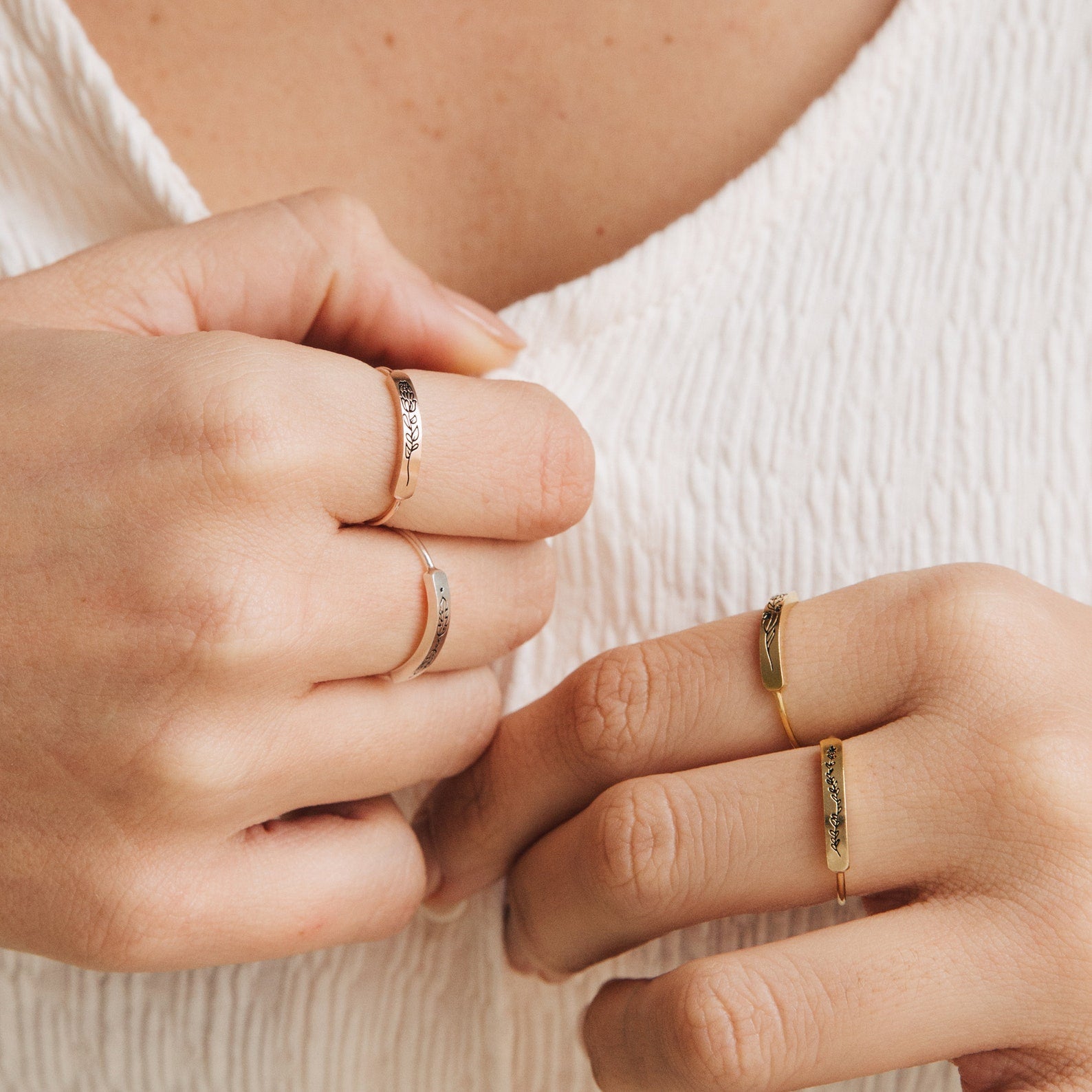 Close-up of hands wearing delicate rings, including a skinny band and the Flower Bar Ring in Sterling Silver, against a textured white fabric background.