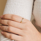 A hand with nude nails wears the Flower Bar Ring in Rose Gold, a delicate skinny band with leaf engravings, resting on a white textured sleeve—an elegant piece of botanical jewelry.