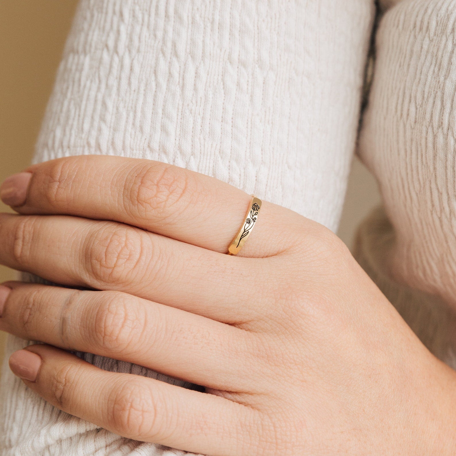 A hand with nude nails wears the Flower Bar Ring in Rose Gold, a delicate skinny band with leaf engravings, resting on a white textured sleeve—an elegant piece of botanical jewelry.