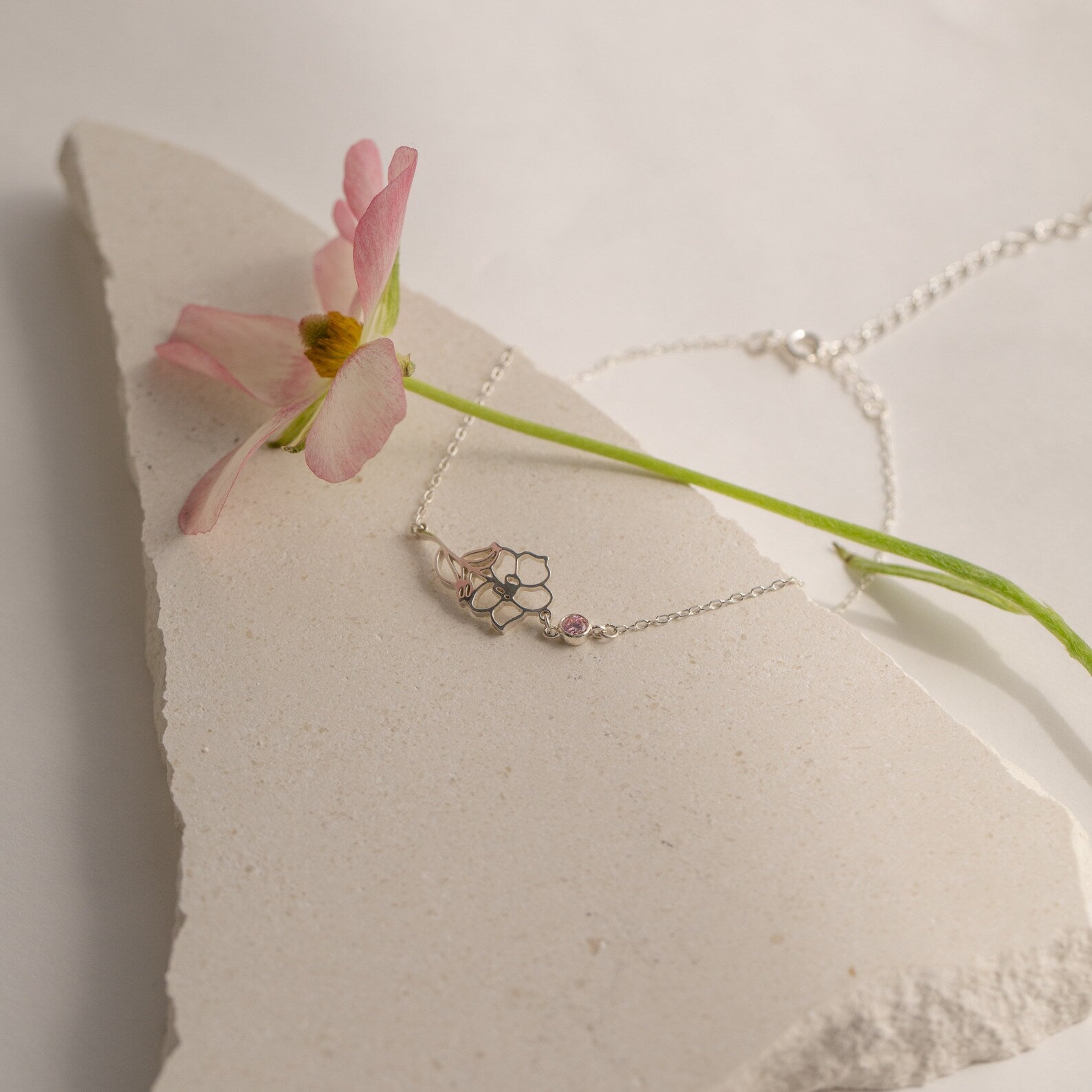 Delicate silver necklace with floral pendant, shown on a stone slab next to a light pink flower—pairs beautifully with the Flower Birthstone Bracelet for a thoughtful birthday gift.