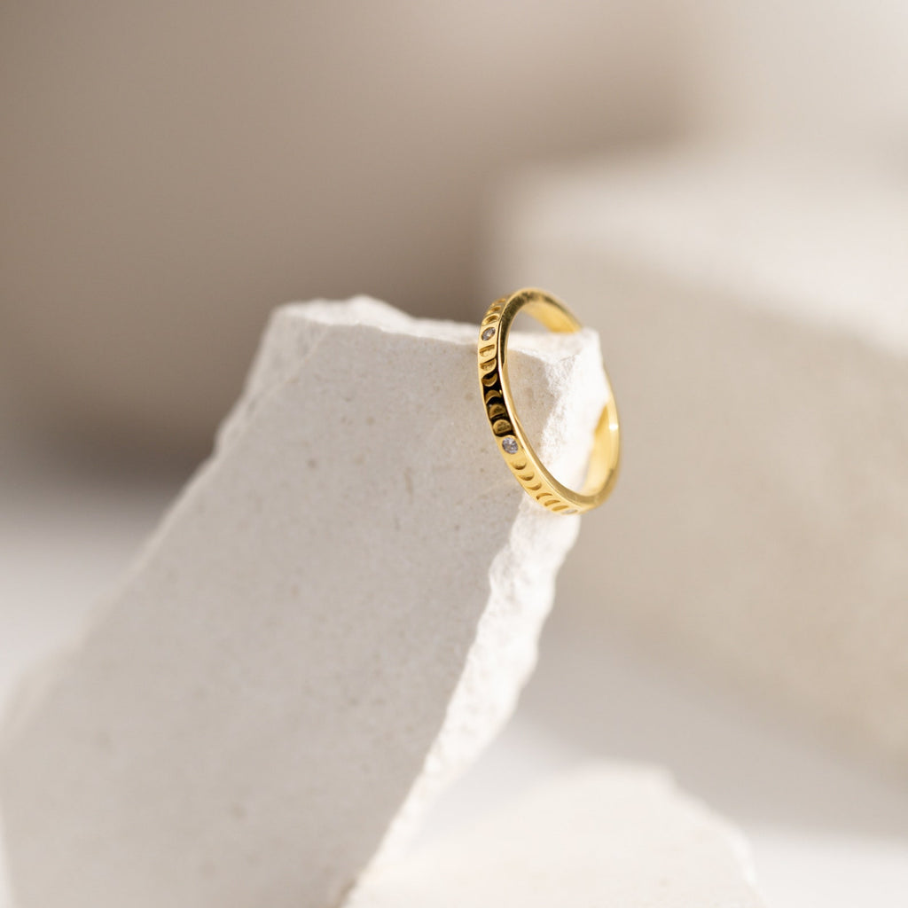 The Augustine Moon Phase Ring, featuring engraved celestial details, is showcased on a white stone against a soft neutral background—ideal for lovers of minimalist moon phase jewelry.