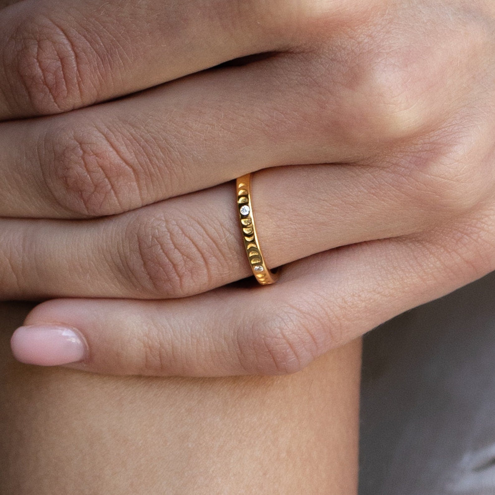 A hand wears the Augustine Moon Phase Ring, a celestial minimalist jewelry piece, and rests gently on another hand with manicured nails.