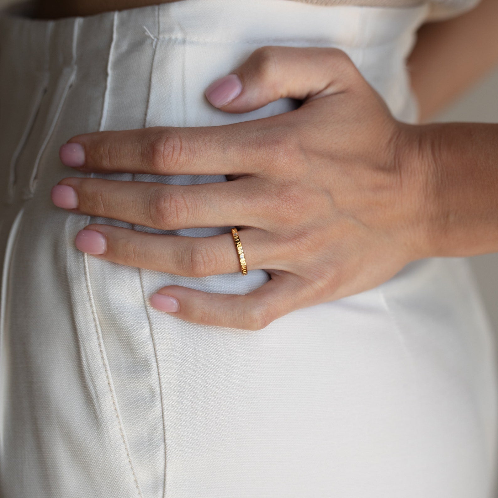 A hand adorned with the Augustine Moon Phase Ring rests on the hip of someone in white high-waisted pants.
