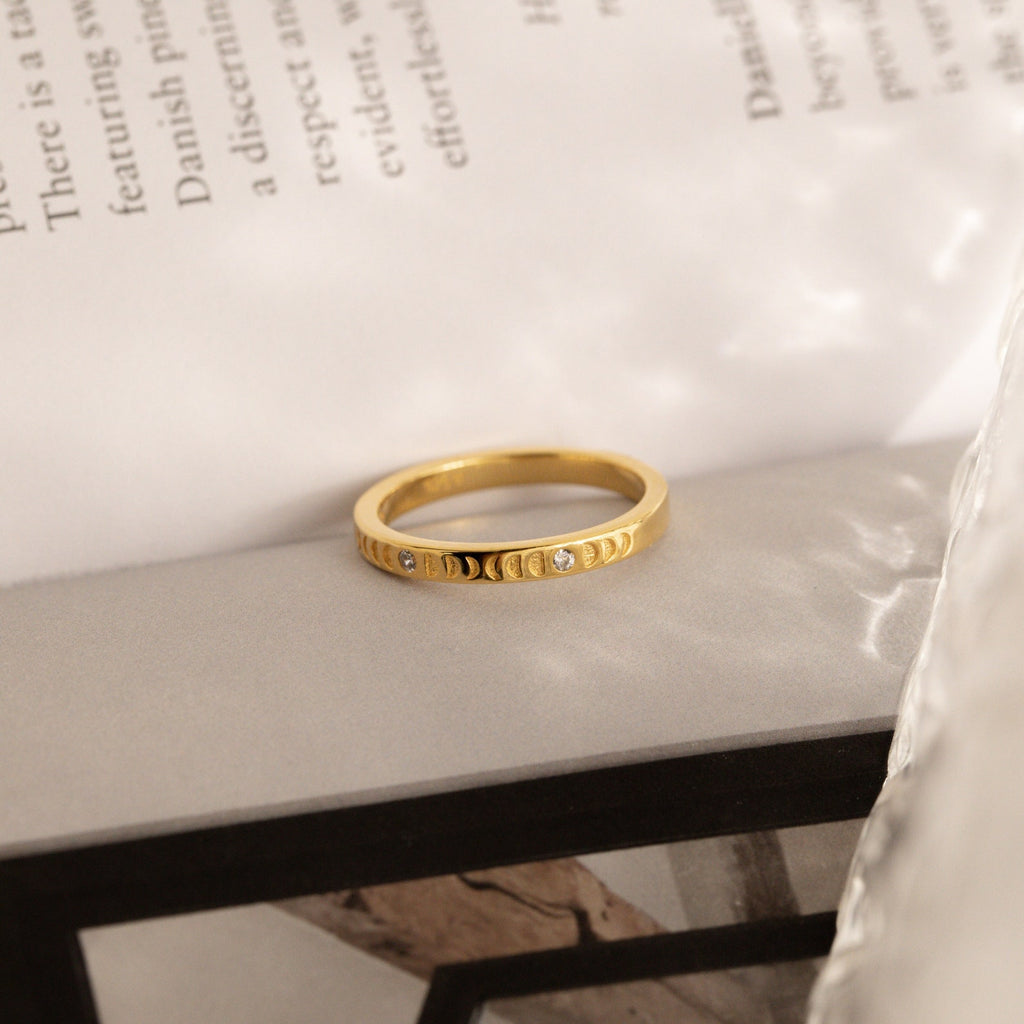 The Augustine Moon Phase Ring, a minimalist piece adorned with small diamonds, rests on a white surface with blurred book text in the background.