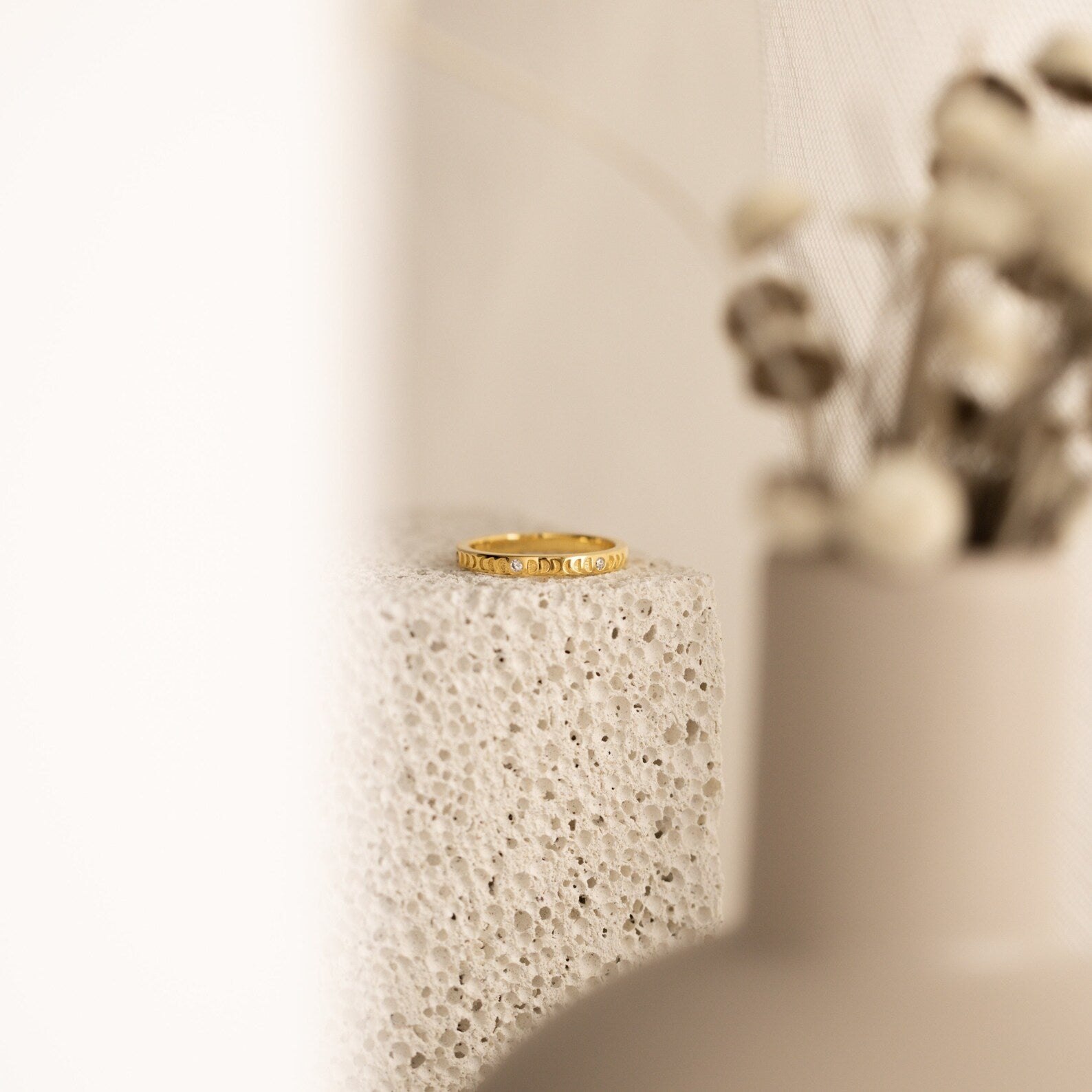 The Augustine Moon Phase Ring rests on a textured stone surface, with blurred dried flowers in the foreground.