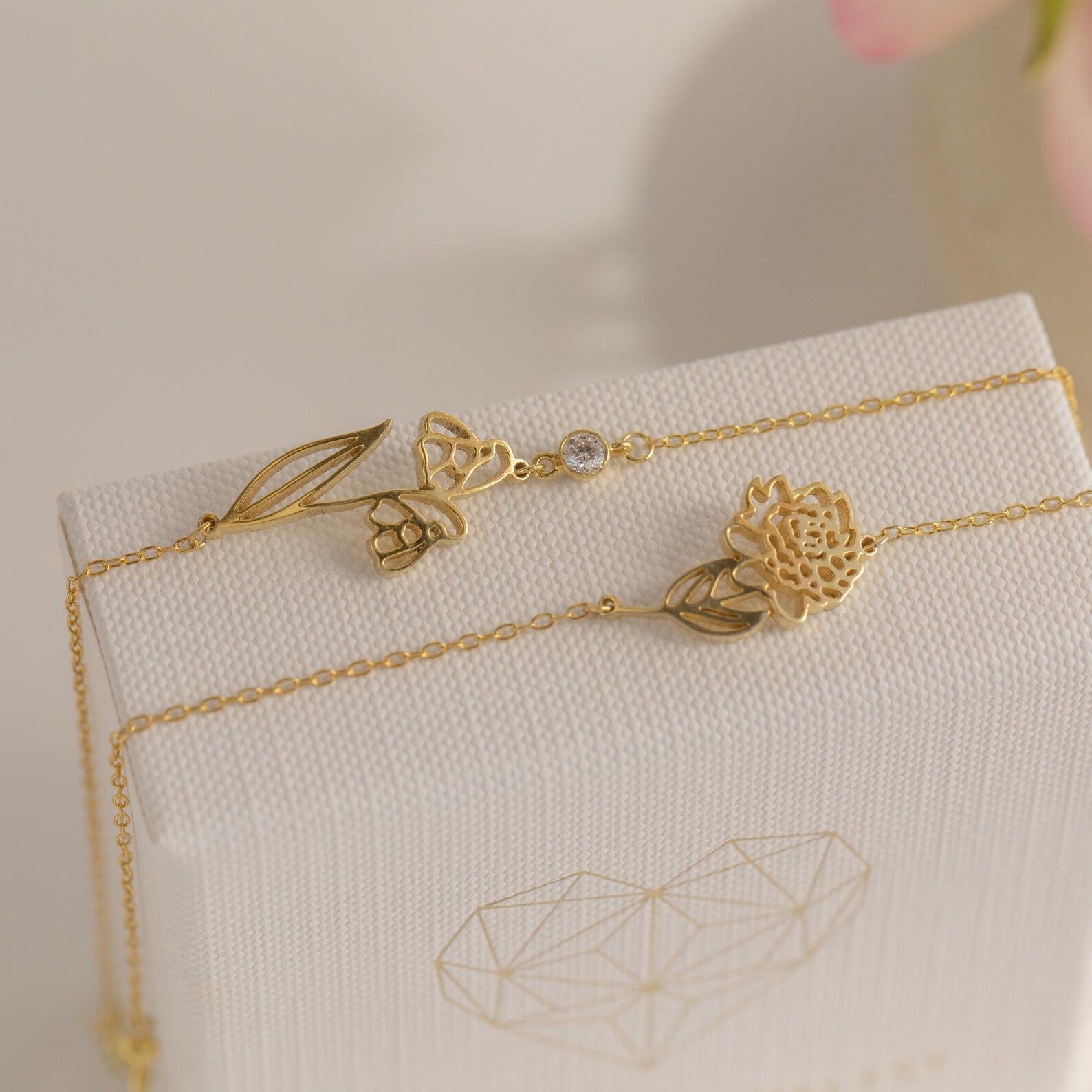 The Custom Flower Bracelet, a delicate gold piece with birth flower and floral charms, is displayed on a white textured box with a geometric heart—an ideal personalized graduation gift.