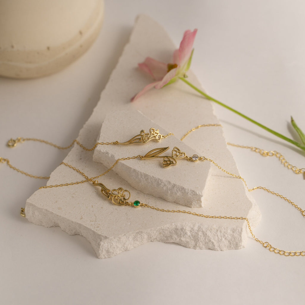 The Custom Flower Bracelet, a delicate gold charm bracelet featuring a birth flower, is displayed on white stone with a pink flower nearby—making it the perfect personalized graduation gift.