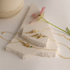 The Custom Flower Bracelet, a delicate gold charm bracelet featuring a birth flower, is displayed on white stone with a pink flower nearby—making it the perfect personalized graduation gift.