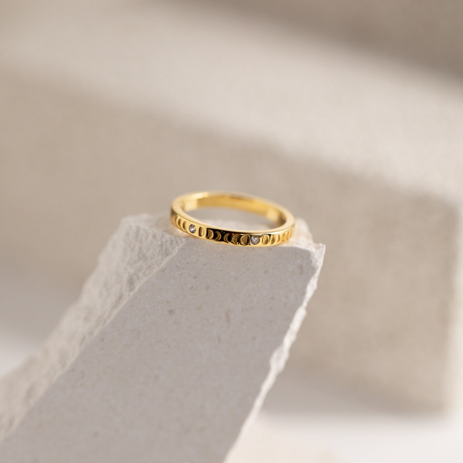 The Augustine Moon Phase Ring features elegant crescent designs and is shown on a textured, light stone surface. This minimalist piece is perfect for anyone who loves celestial-inspired jewelry.