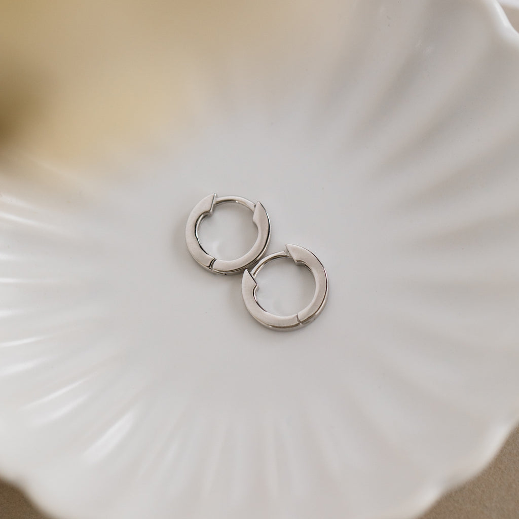 The Editor III Huggies, a pair of silver hoop earrings, are displayed on a white, scallop-shaped ceramic dish, highlighting their minimalist elegance.