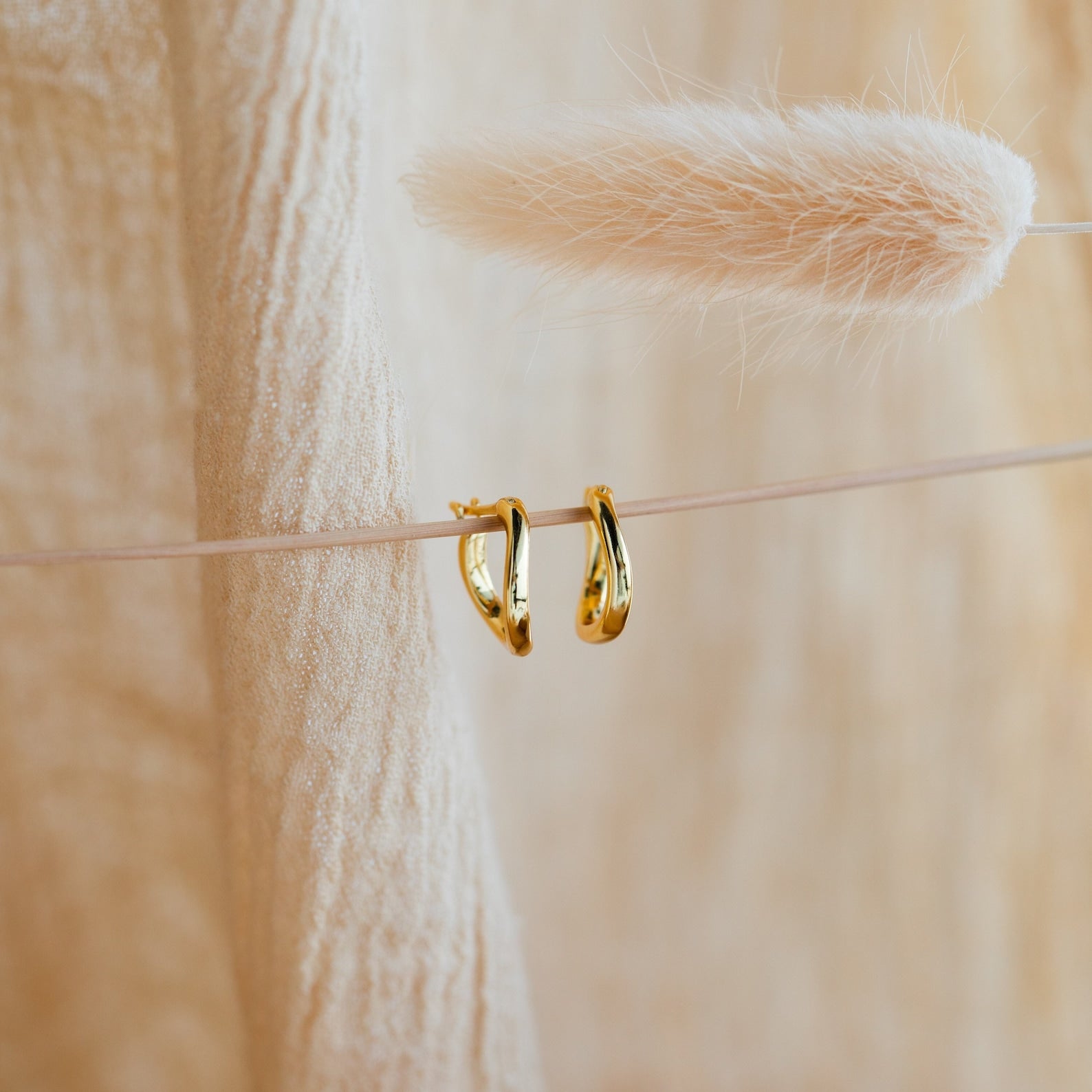 The McKenna Abstract Hoops feature an organic gold design, displayed against soft beige fabric and dried grass—perfect for elevating your everyday look with a touch of unique elegance.