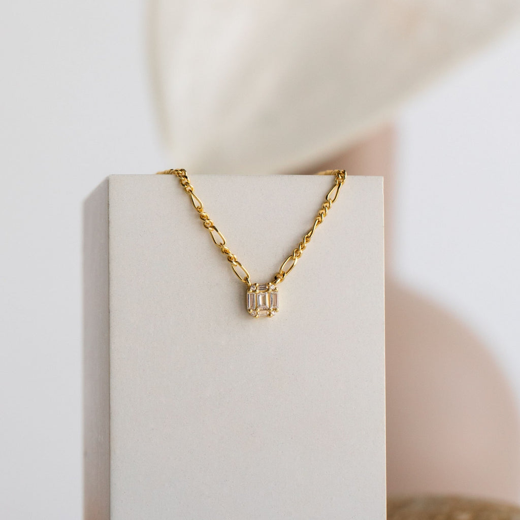 Gold necklace with a bold twisted chain and rectangular pendant featuring three vertically-set baguette-cut stones, displayed draped over a soft beige jewelry stand.
