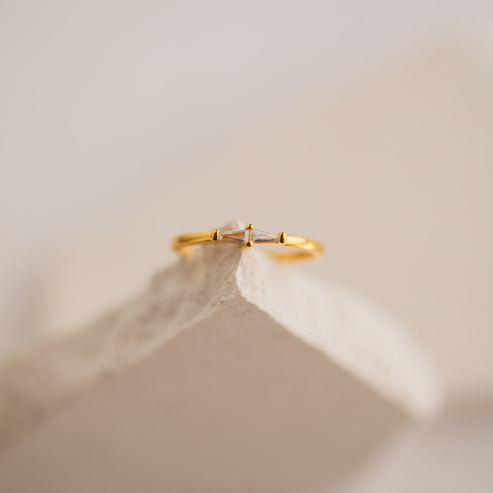The Eleanor Baguette Diamond Ring, a delicate gold band featuring a baguette diamond, is displayed on a textured, light-colored stone surface.