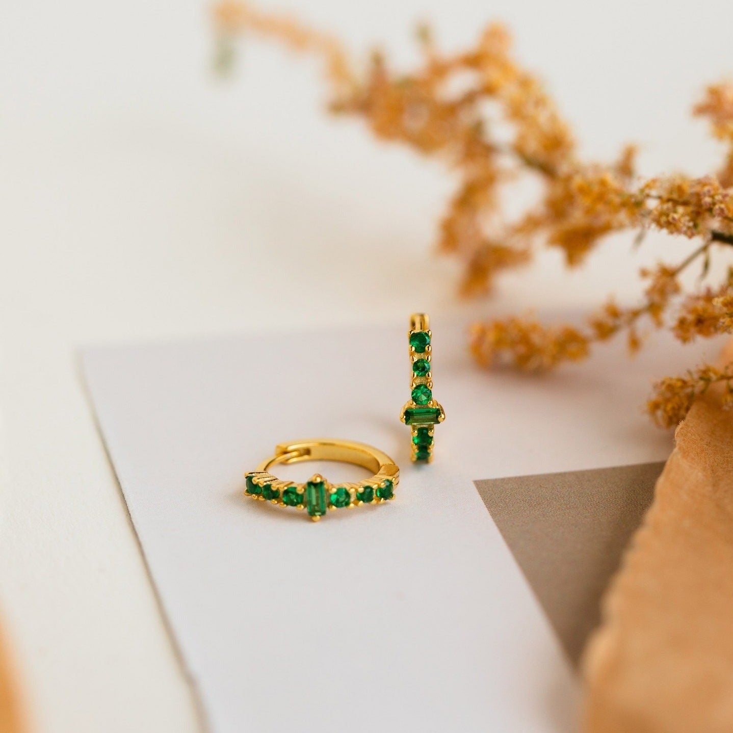 Ireland Emerald Huggies gold earrings with green gemstones shown on a white surface, accented by dried flowers in the background.