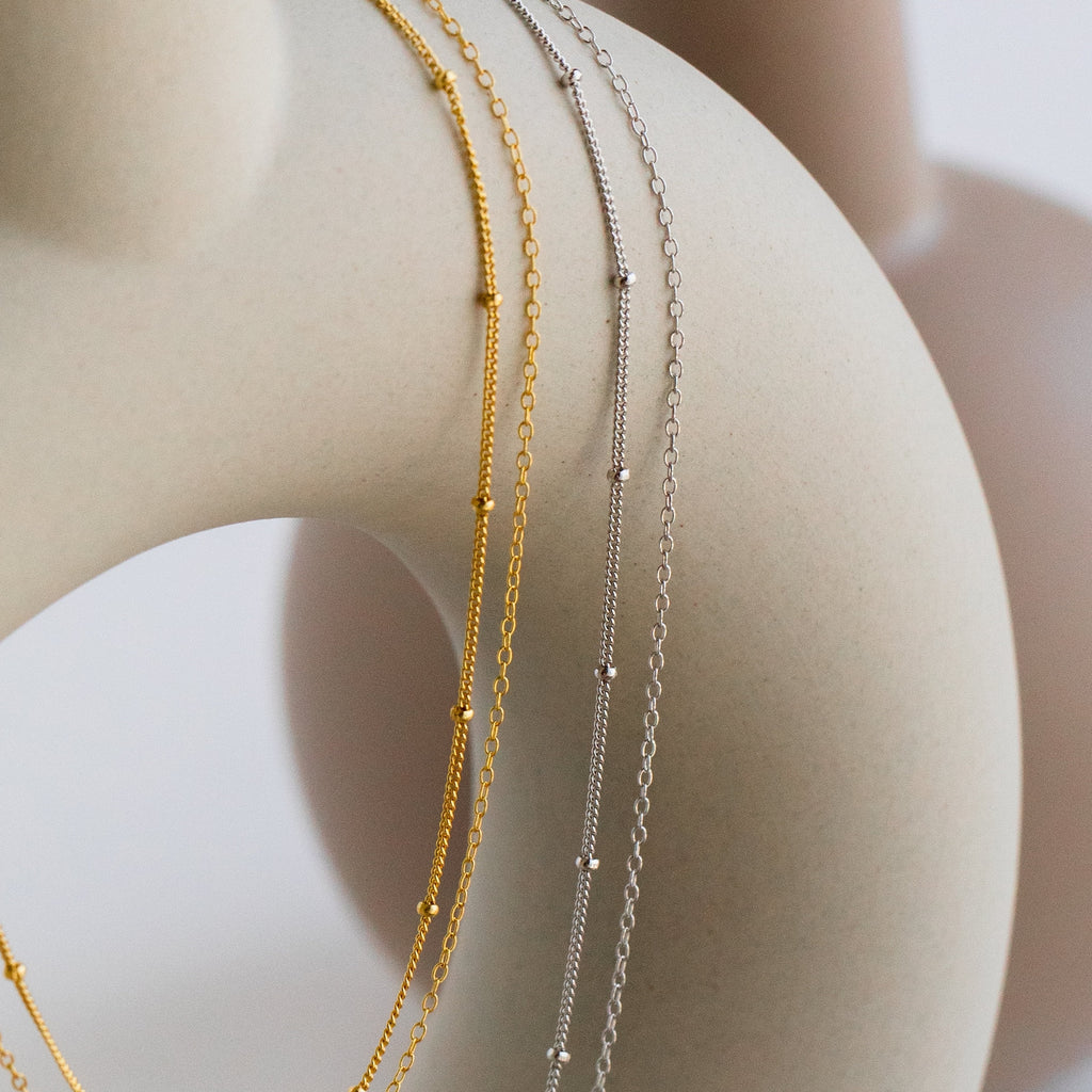 Four delicate gold and silver chains, including the minimalist Beaded Duo Chain Necklace, are elegantly draped over a sleek, abstract beige sculpture.