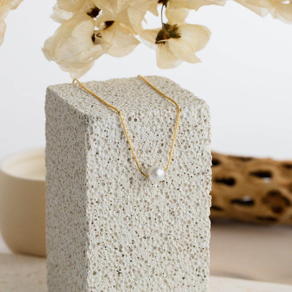 The Kennedy Pearl Necklace is shown on a textured stone block with dried flowers behind it—an ideal pick for minimalist style or as elegant wedding jewelry.