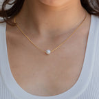 Woman wearing a minimalist gold chain necklace with a single round white pearl centered at the collarbone, styled with a white ribbed scoop-neck tank.