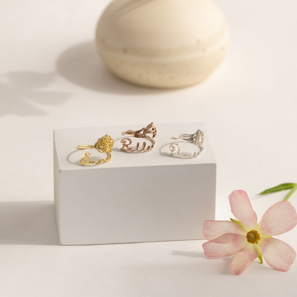 Four Flower Name Rings displayed on a white block, with a pink flower and ceramic accent—perfect as personalized flower jewelry or a custom Birth Flower Name Ring, making them an ideal gift for mom.
