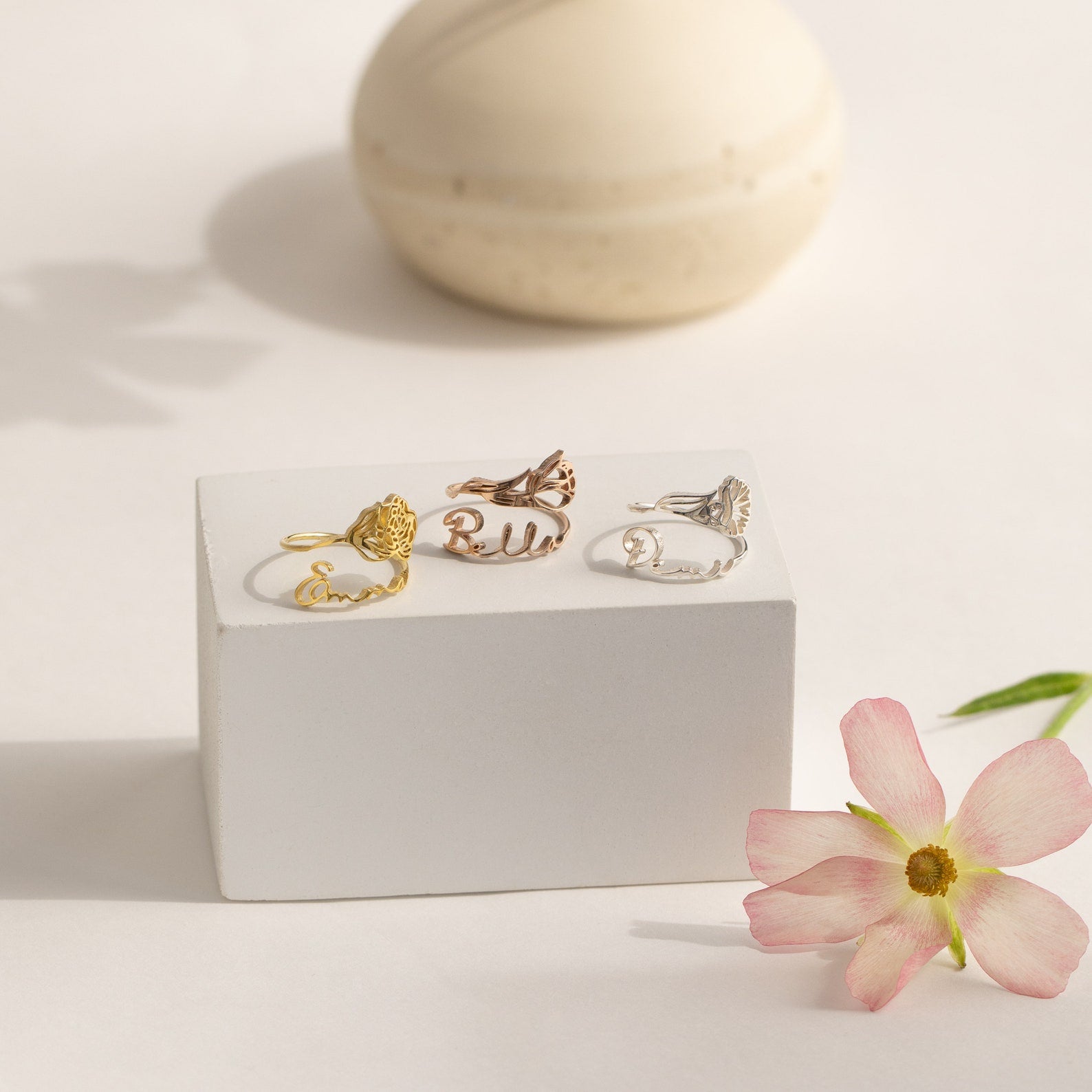 Four Flower Name Rings displayed on a white block, with a pink flower and ceramic accent—perfect as personalized flower jewelry or a custom Birth Flower Name Ring, making them an ideal gift for mom.