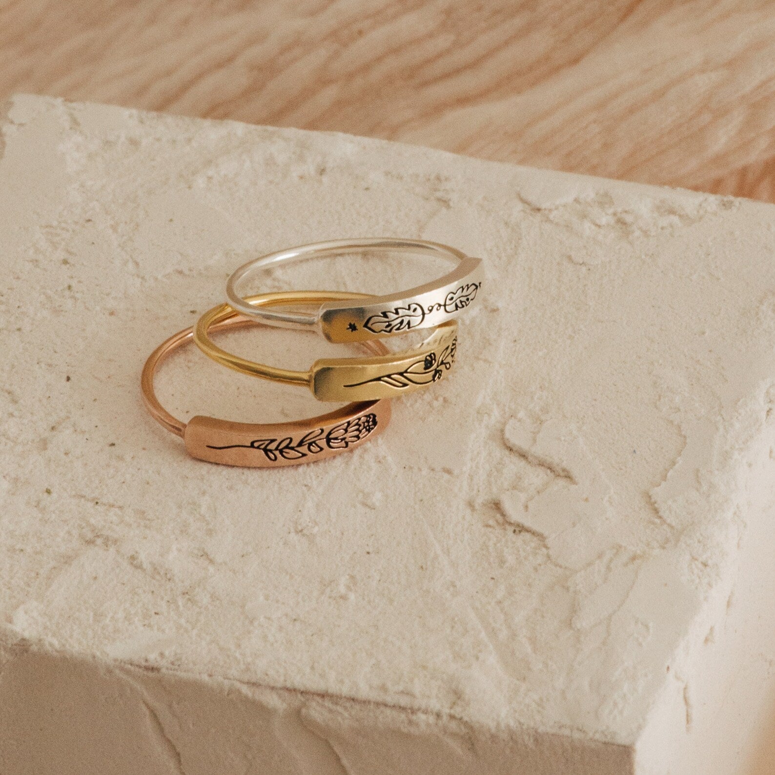 The Flower Bar Ring in Rose Gold features a sleek metal band with engraved botanical designs, stacked on a textured, light-colored stone surface—an ideal choice for lovers of nature-inspired jewelry.