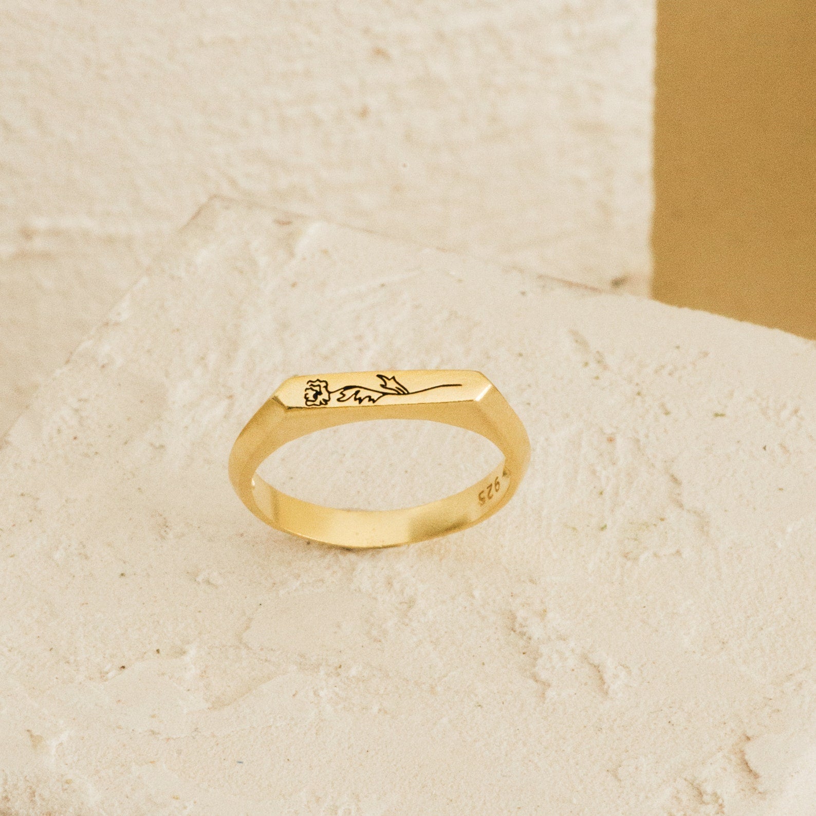 The Thin Flower Signet Ring features an engraved minimalist flower design on gold, resting on a textured off-white surface—an ideal personalized pinky ring for botanical jewelry enthusiasts.
