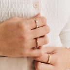 Close-up of hands wearing three engraved rings in gold and silver, including the Thin Flower Signet Ring as a personalized pinky ring, with a textured cream shirt in the background.