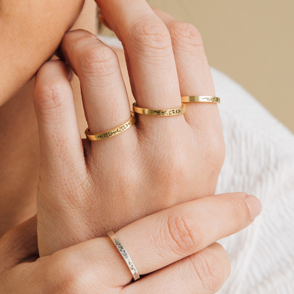 Close-up of a hand wearing four engraved gold rings, including the Stacking Flower Band on the middle or ring finger, a skinny band, botanical jewelry, and a silver ring on the pinky.