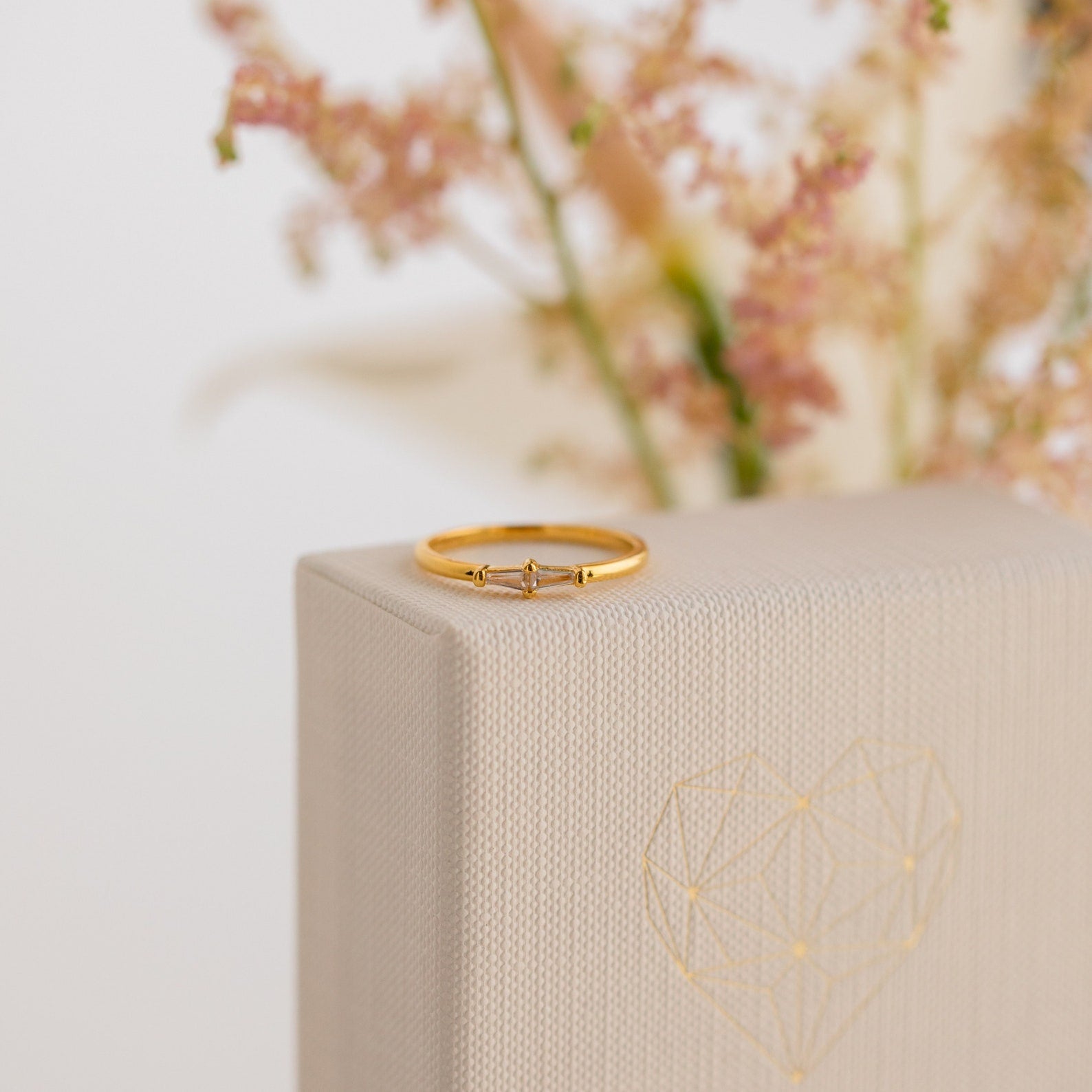 The Eleanor Baguette Diamond Ring rests atop a textured beige box adorned with a geometric heart design, while delicate flowers add a soft background touch.