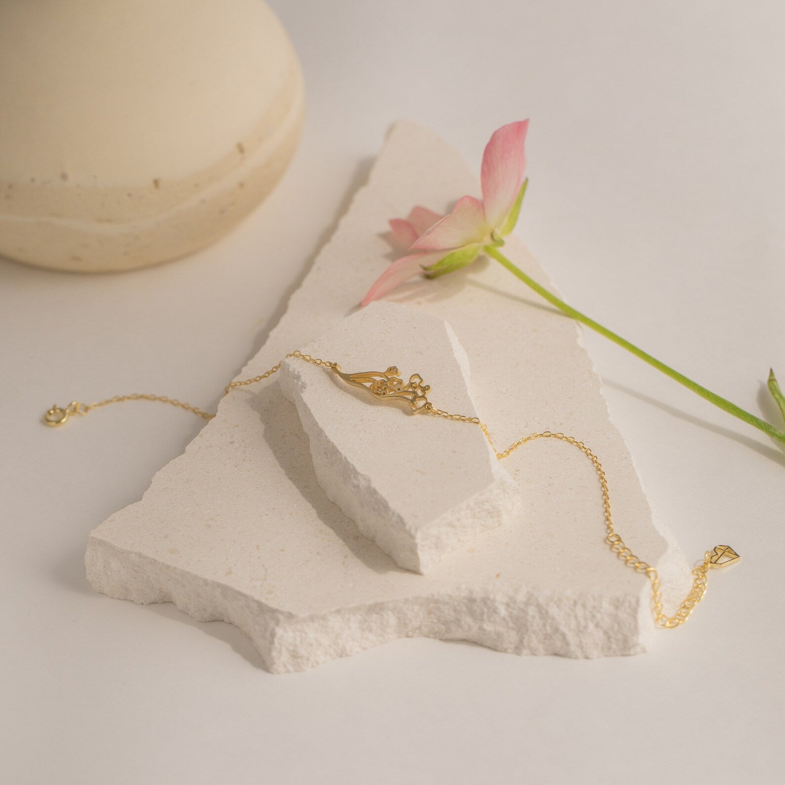 Custom Flower Bracelet in gold, no stone, displayed on textured white stone with a pink birth flower and a cream vase in the background; an ideal personalized graduation gift.