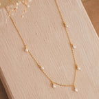 Delicate gold chain necklace with evenly spaced small freshwater pearl drops, displayed on a textured beige surface with dried floral accents.