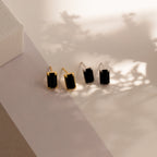 Two pairs of Rectangle Black Onyx Studs—one with gold, one with silver trim—rest on a light surface with soft shadows. These elegant earrings add timeless style to any look.