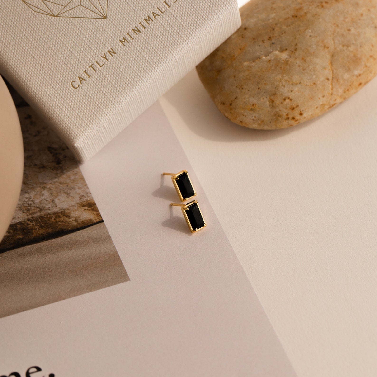 Rectangle Black Onyx Studs featuring gold and rectangular black stones rest on a white surface beside a box, a stone, and printed text, adding a timeless vintage touch.