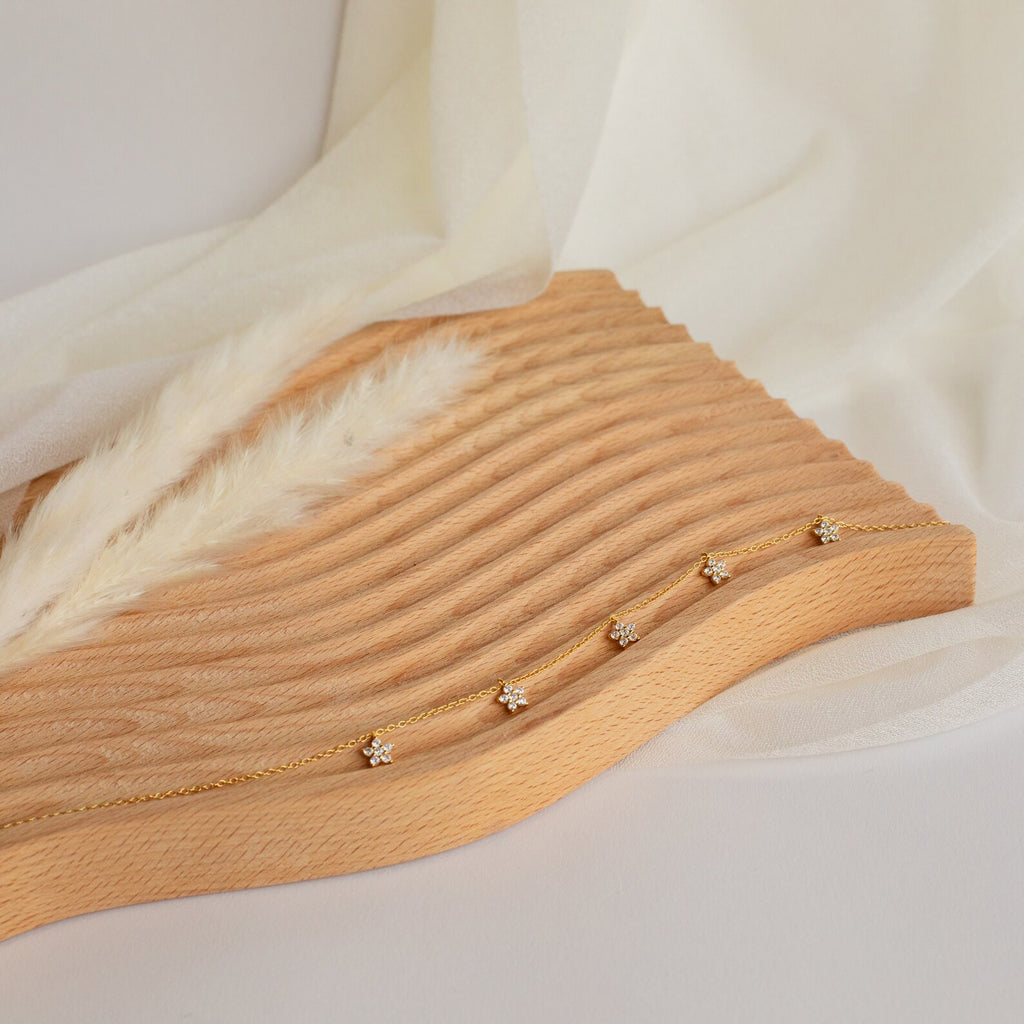 The Nanala Diamond Flower Necklace is displayed elegantly on a wavy wooden board, paired with a gold bracelet, and accented with fabric and dried grass for added charm.