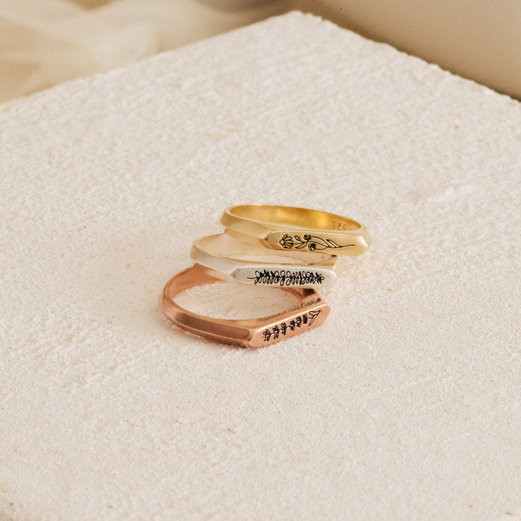 Three engraved Thin Flower Signet Rings—gold, silver, and rose gold—stacked on a textured cream surface. This botanical set features a personalized pinky ring for a delicate, unique touch.