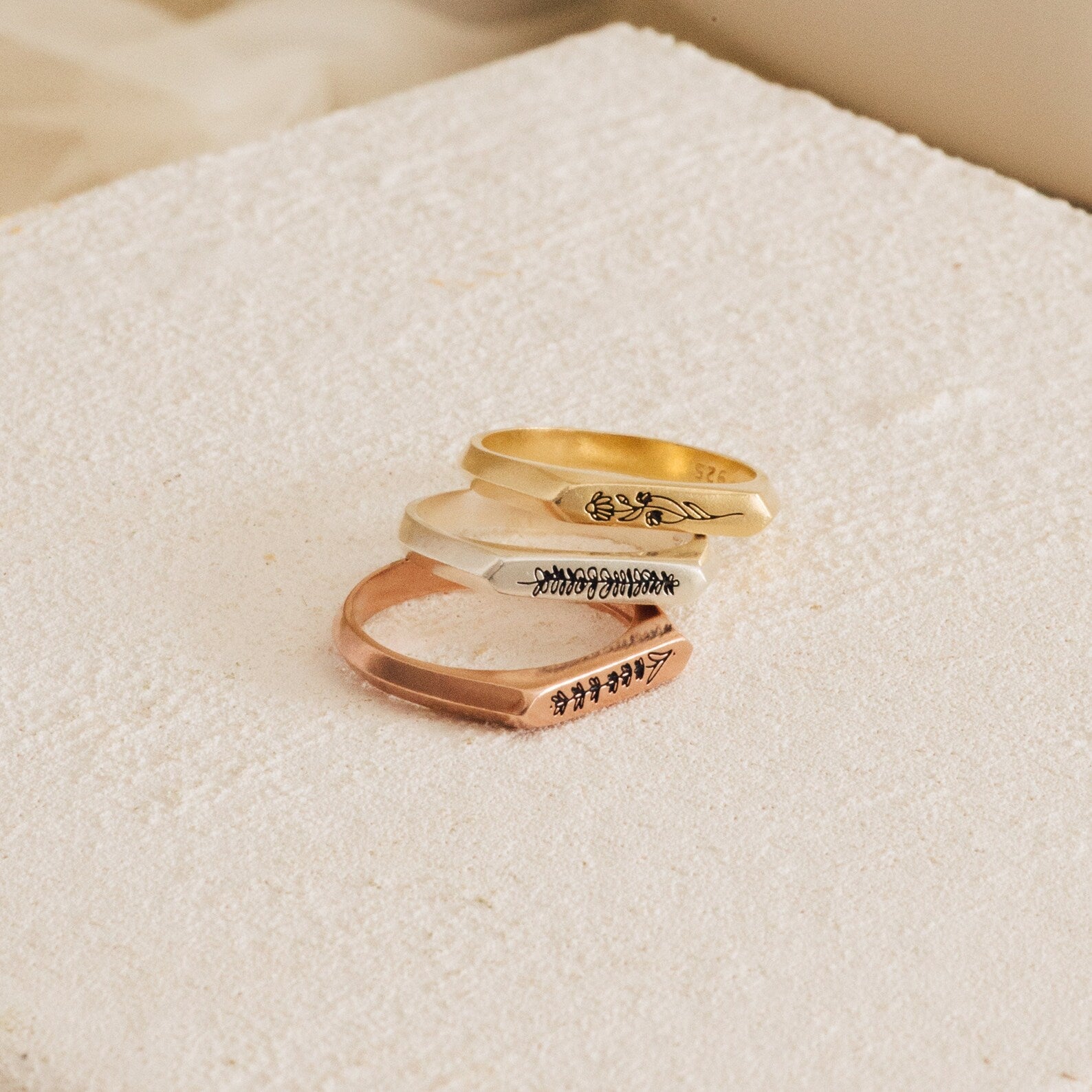 Three engraved Thin Flower Signet Rings—gold, silver, and rose gold—stacked on a textured cream surface. This botanical set features a personalized pinky ring for a delicate, unique touch.