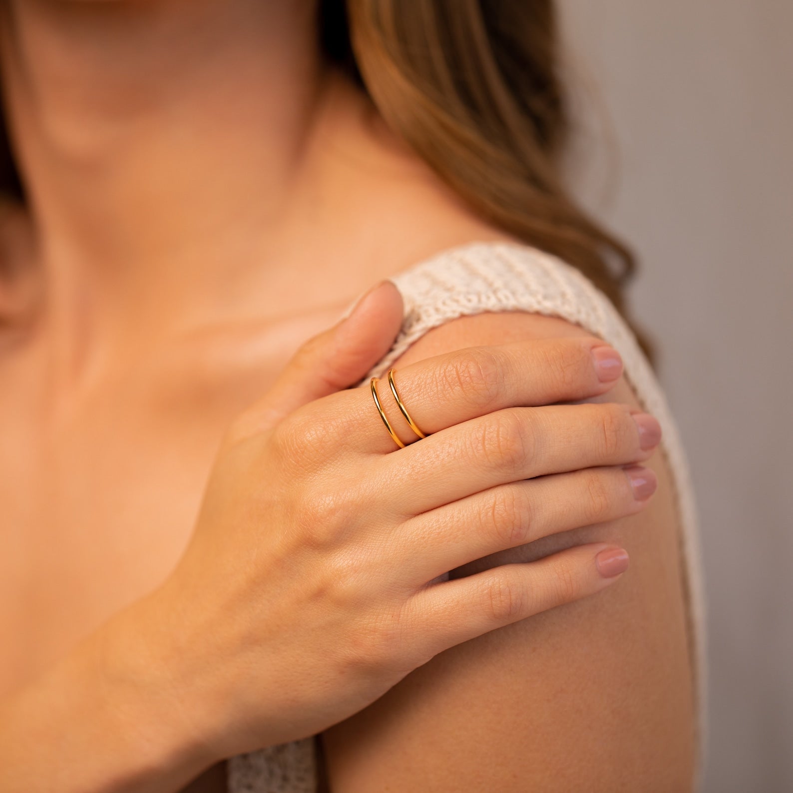 A woman rests her hand on her shoulder, wearing the Everly Thin Duo Band—a simple gold stacking ring with an adjustable band—and light pink nail polish.