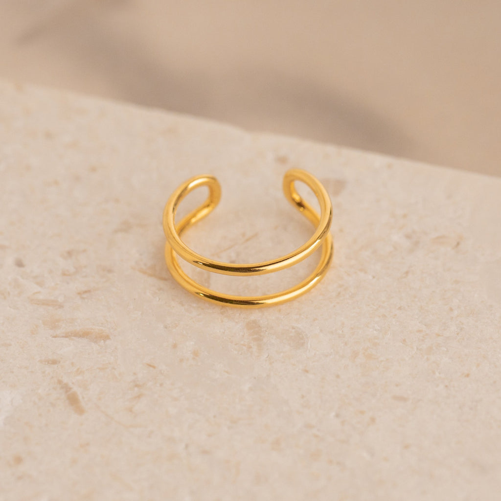 The Everly Thin Duo Band is a gold open cuff ring featuring an adjustable double band design, ideal for stacking. Shown on a textured beige stone surface.