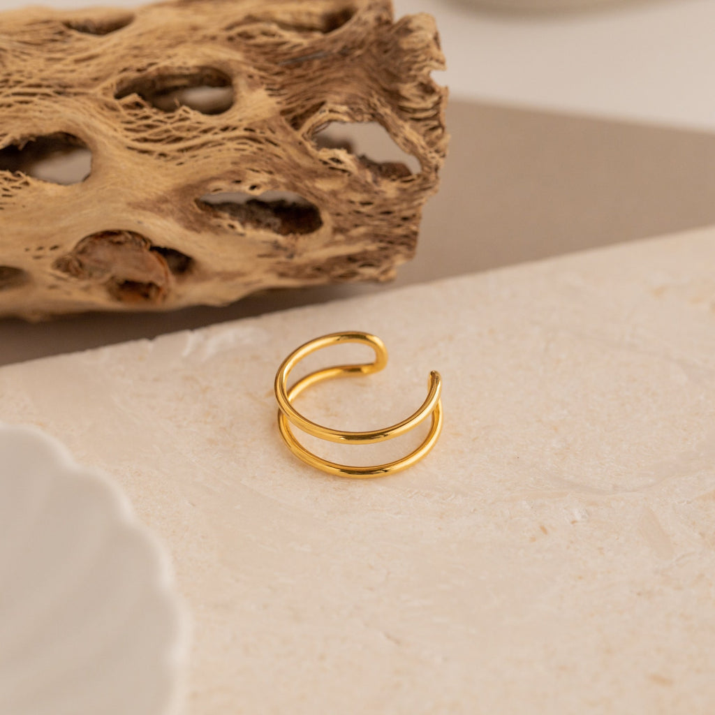 The Everly Thin Duo Band sits on a beige textured surface with a dried natural accent in the background, showcasing its elegant double-band design—ideal as an adjustable stacking ring.