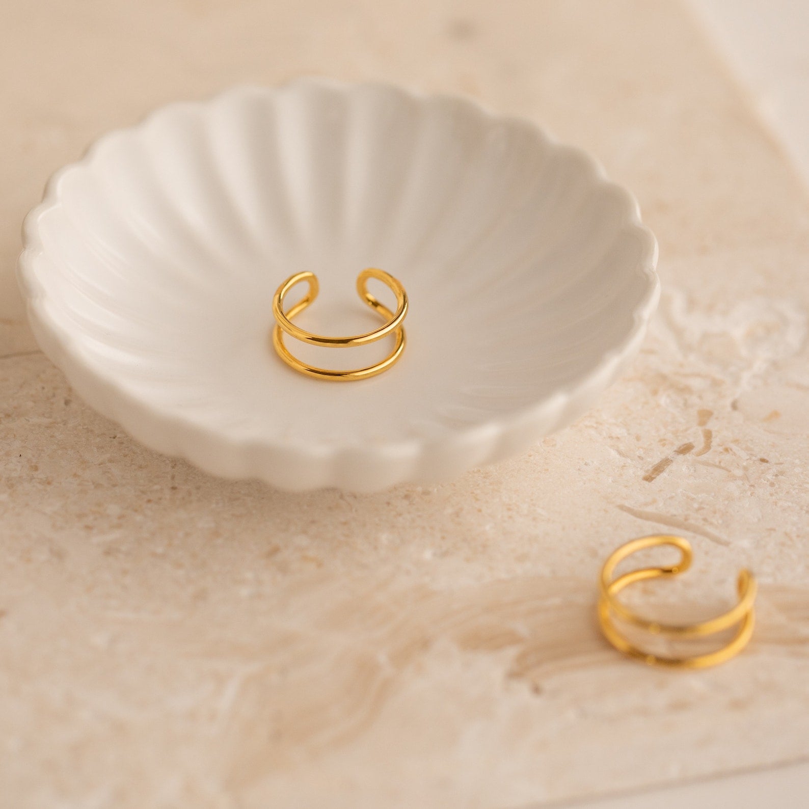 An Everly Thin Duo Band, a gold adjustable ring, rests in a scalloped white dish, while another stacking ring is displayed on a beige textured surface nearby.