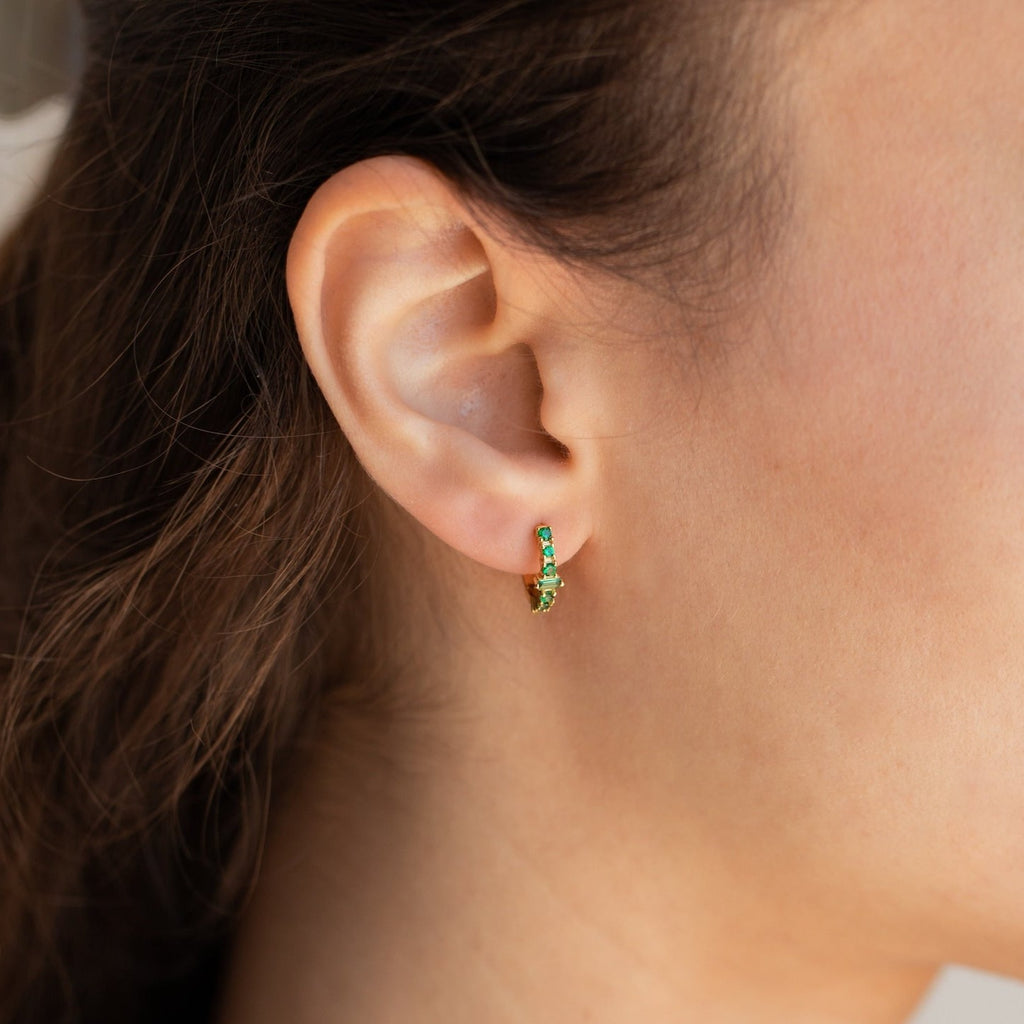 Close-up of a woman's ear wearing the Ireland Emerald Huggies, small gold hoop earrings featuring brilliant green stones.
