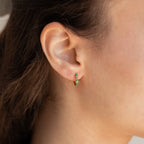 Close-up of a woman's ear wearing the Ireland Emerald Huggies, small gold hoop earrings featuring brilliant green stones.