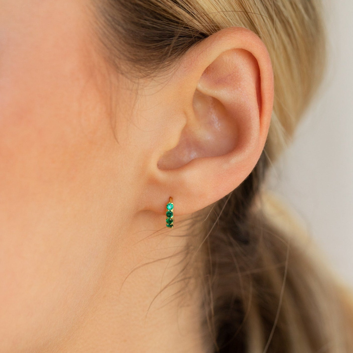 Close-up of a woman's ear wearing the Elise Emerald Huggies—small gold earrings adorned with green gemstones, ideal for fans of elegant green jewelry.