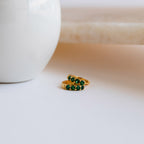 The Elise Emerald Huggies, two gold rings set with emerald stones, rest on a white surface beside a white ceramic object, bringing an elegant pop of green to the scene.