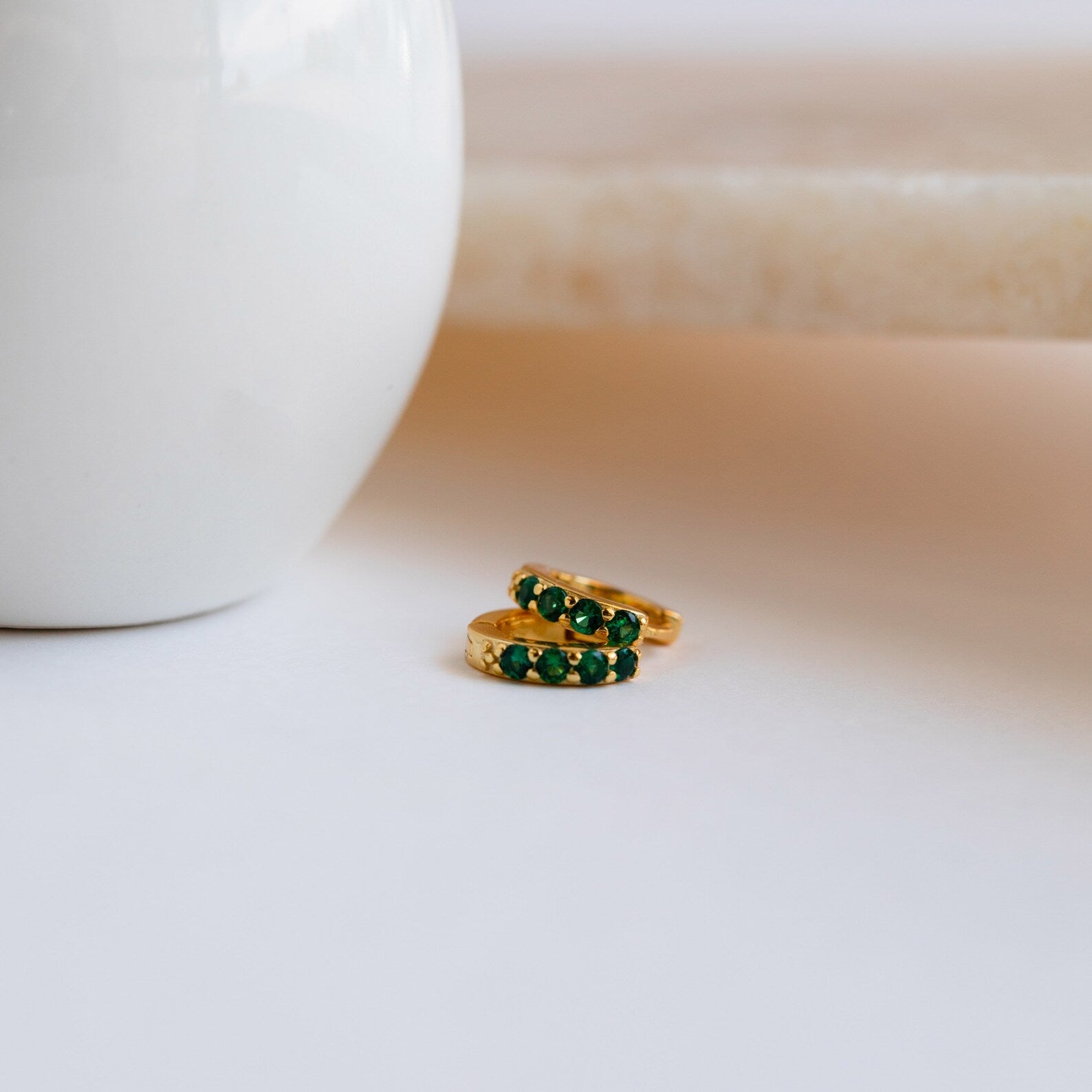 The Elise Emerald Huggies, two gold rings set with emerald stones, rest on a white surface beside a white ceramic object, bringing an elegant pop of green to the scene.