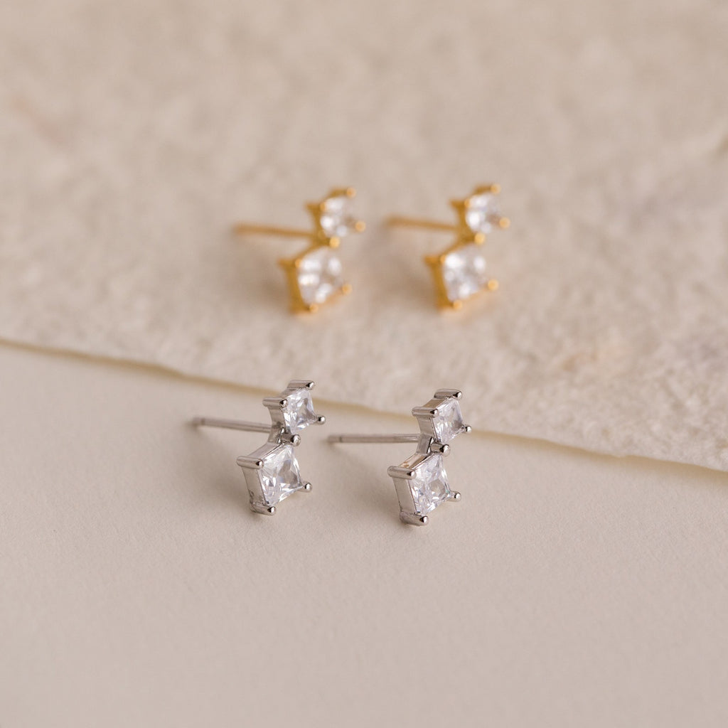 The Aria Diamond Drop Studs set includes two pairs of lightweight stud earrings—one gold, one silver—each featuring two square-cut clear stones. Perfect for any occasion, they’re beautifully displayed on a neutral background.