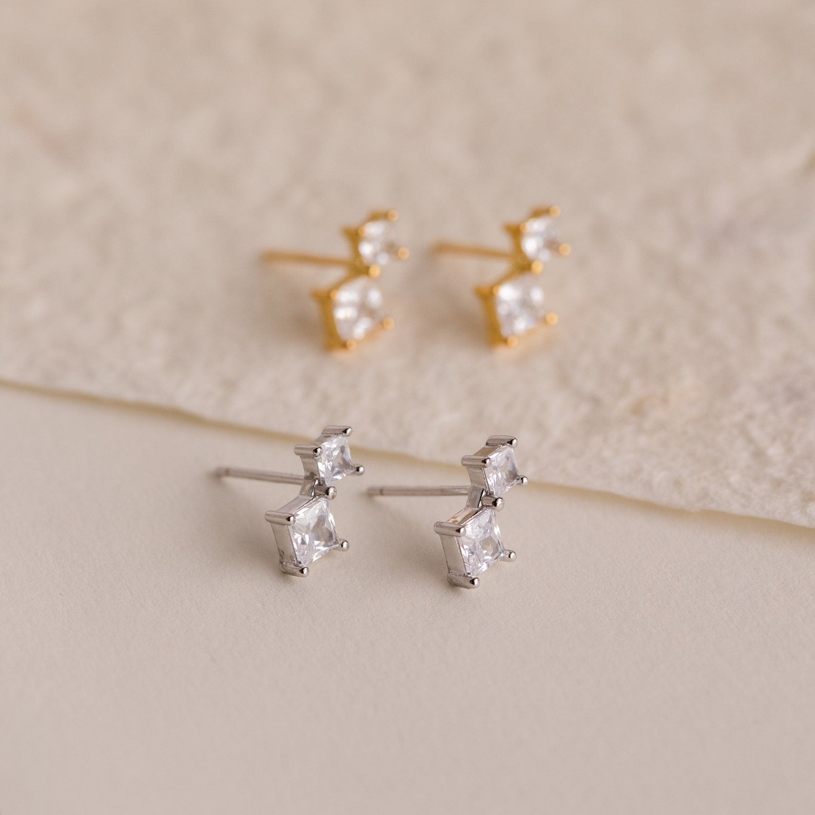 The Aria Diamond Drop Studs set includes two pairs of lightweight stud earrings—one gold, one silver—each featuring two square-cut clear stones. Perfect for any occasion, they’re beautifully displayed on a neutral background.