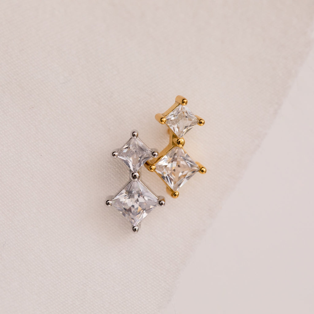 Aria Diamond Drop Studs featuring gold and silver crystal with three square gemstones, displayed on a light fabric background.